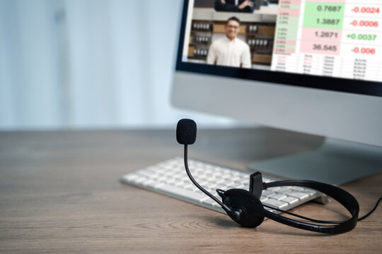 Work from home setup with headset on desk, showing person in online video conference on desktop monitor with financial charts, representing remote communication and digital teamwork efficiency. - Powered by Adobe