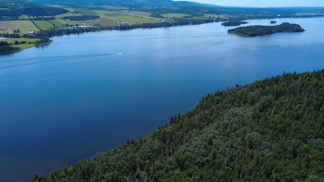 Panoramic aerial view of the islands of a vast blue freshwater lake where the wake of a solitary boat disturbs the calm water in a rural agricultural and forest setting. Lake Matapedia, Quebec, Canada