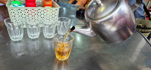 Lift the stainless steel kettle to pour tea into the glasses. Four empty glasses are placed in a...