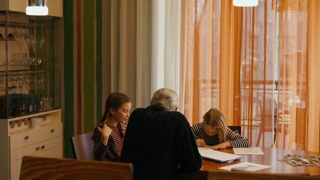 Grandpa approaches and sits at a table where two granddaughters are sitting and drawing, one of whom is talking
