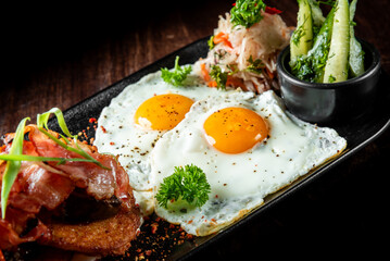 Delicious breakfast plate with sunny-side-up eggs, crispy bacon on toast, coleslaw, and pickled cucumbers, garnished with herbs on a dark background.