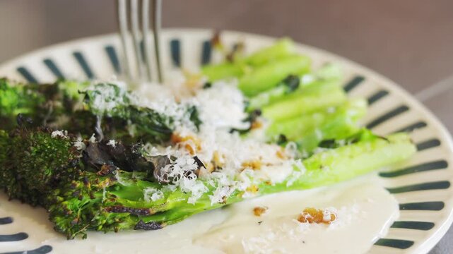 Close-up of roasted broccolini with parmesan and crunchy crumbs on ceramic plate. Selective focus. Fork pressing vegetables. Healthy, vegetarian, flavorful dish captured in natural warm lighting.