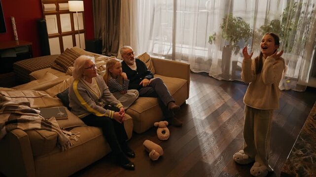 Grandfather, grandmother, and granddaughter are sitting on the couch and guessing what her sister, who is standing across from them, is showing