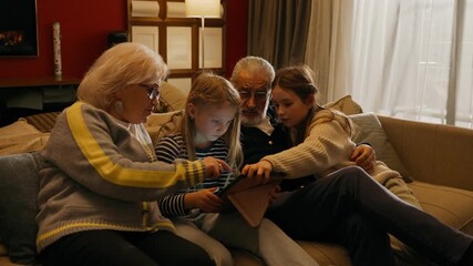 Two granddaughters and a grandmother clicking on a computer tablet while sitting on the couch with their grandfather - Powered by Adobe