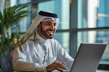 An Arab man in traditional dress smiles while sitting at a laptop.