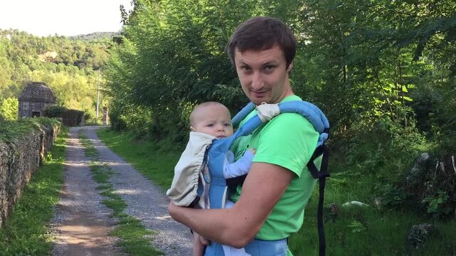 Father carries baby in carrier and talks to him while walking along scenic outdoor path. The baby looks curiously at camera, then child hides his face, creating a tender family moment amidst nature