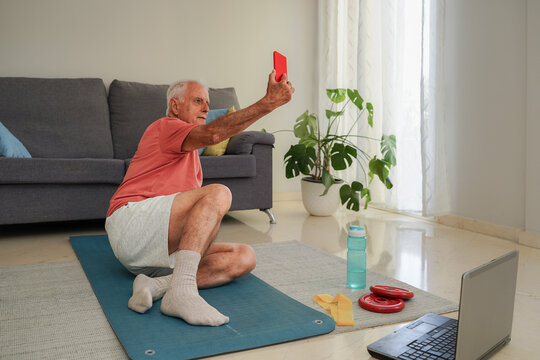 Senior man taking selfie while exercising at home with laptop and smartphone - Powered by Adobe