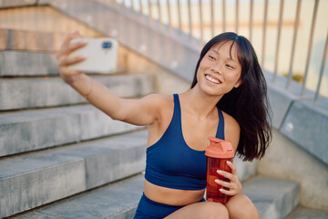 Smiling asian sportswoman taking selfie with smartphone while holding shaker sitting on urban stairs