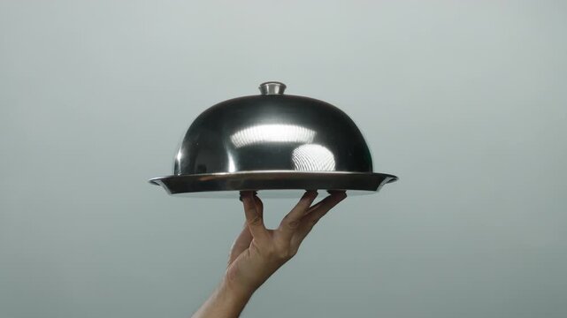 Hand holding silver cloche against white background, emphasizing service and presentation in a minimalist context.