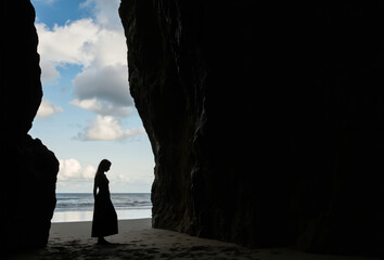 Woman stands alone on sandy beach, facing rocky cliff. Black dress, clear blue sky with scattered clouds. Low angle shot emphasizes cliff height and vast sky.