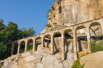 Perspective view of stone arcade stepping along a rocky slope with repeating arches in soft evening...