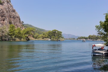 A scenic landscape photo shows a wide river with mountains rising on the left, under a clear blue sky in Dalyan - Ortaca - Mugla - Turkey