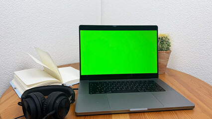 Laptop with green chroma key screen on wooden desk, modern workspace setup with cactus plant, coffee cup, books and headphones, technology background for mockup, isolated computer screen for editing