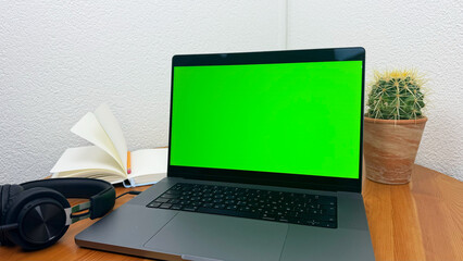 Laptop with green chroma key screen on wooden desk, modern workspace setup with cactus plant, coffee cup, books and headphones, technology background for mockup, isolated computer screen for editing