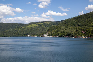 A tranquil lake is nestled amidst lush green hills and a dense forest under a bright blue sky with fluffy clouds.