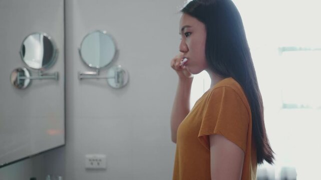 A young woman in a yellow top stands in front of a bathroom mirror, thoughtfully examining her reflection. The atmosphere is calm and serene, with a focus on self-care and personal reflection in a bri