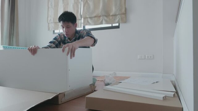A young man is focused on assembling furniture in a well-lit room. The space is organized with tools and materials around him, highlighting a DIY home improvement project.