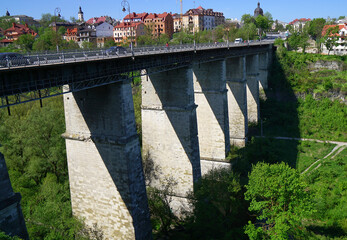A wide shot of a historic stone bridge with tall pillars spanning a lush green gorge, with a...