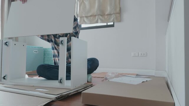 A person is assembling a white furniture item in a bright room. Tools and materials are scattered around, showcasing a typical DIY furniture project. The workspace is organized with an attention to de