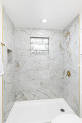 A walk-in shower with marble tiled walls, gold shower head, and glass block window.