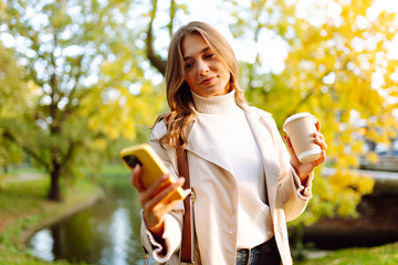Portrait of a young woman with a phone and a cup of coffee walking in an autumn park on sunny day. Beautiful tourist enjoying sunset and looking at the screen of mobile. Walking concept. Lifestyle.