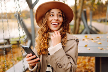 A beautiful woman in casual clothes and a phone enjoys an autumn park at sunset. A woman in a hat...