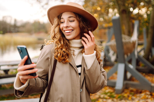 A beautiful woman in casual clothes and a phone enjoys an autumn park at sunset. A woman in a hat is texting on her phone while riding a swing outdoors. Autumn walk.