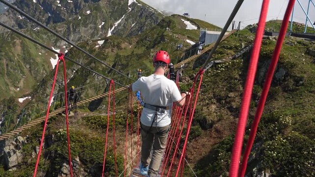 A young man wearing a helmet walks across a suspension bridge in the mountains between two peaks, an extreme attraction for travelers, Rosa Khutor.