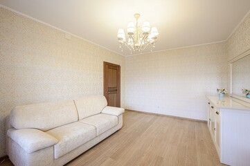 cozy room with a beige sofa, patterned wallpaper, a wooden door, and a white cabinet. Chandelier on the ceiling and light wood floors