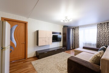 Cozy living room with a beige sofa, brown furniture, a TV set, and a fluffy rug. The room is well-lit with a window and a ceiling light