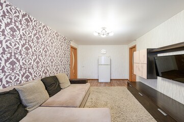 Cozy living room with a beige sofa, TV, and patterned wallpaper. The room includes a fridge and wardrobe