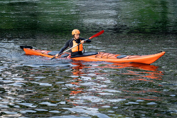 A man wearing an orange helmet and life vest is paddling an orange kayak on a river. He uses a red...
