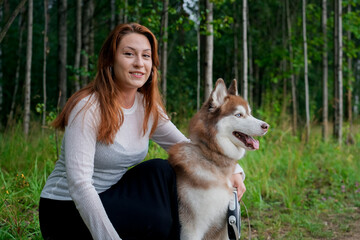 A smiling woman with auburn hair is walking in the forest with her beautiful red and white Siberian...