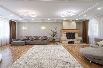 Bright living room with a gray sectional sofa, patterned rug, and stone fireplace. Natural light filters through sheer curtains. Two modern chandeliers illuminate the space