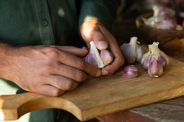 Close-up image of male hands peeling a garlic bulb on a rustic wooden cutting board, symbolizing fresh cooking preparation, natural ingredients, and farm-to-table culinary process