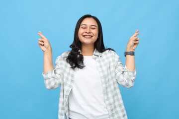 Young Asian woman smiling in casual clothes, crossing fingers with closed eyes, making a wish or...