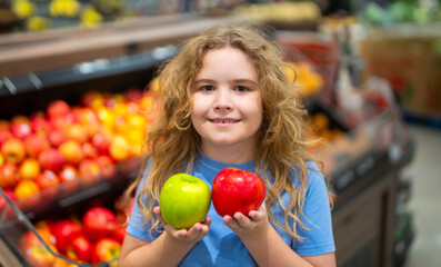 Fototapeta premium Kid holding a fresh apple in fruit store. Cute child holdi apple in fruit market. Red and green apples in kids hand. Apples in kids hand. Child face close-up. Child choosing a apple.