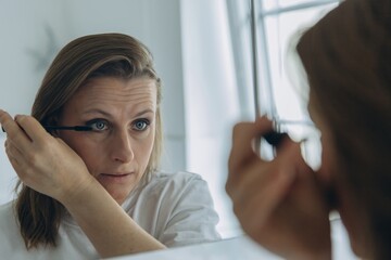Middle-aged woman applying eye makeup with a brush in front of a bathroom mirror. Beauty routine...