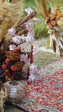 Vertical video. Decor near the wedding arch. Boho style ceremony on a carpet outside in the park. Pastel flower arrangements next to the arch.