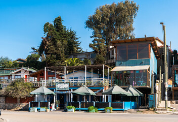 Beachfront restaurant and wooden houses on the Chilean coast under clear blue sky. 
Scenic view of a seaside restaurant with outdoor terraces, umbrellas, and wooden houses on a hillside.