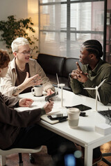 Three diverse gen Z male and female colleagues sitting at office desk with turbine models and solar panel gadgets on it, discussing eco-friendly technologies