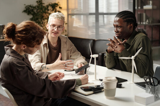 Group of three diverse gen Z male and female colleagues sitting at office desk with turbine models on it, discussing eco-friendly technologies