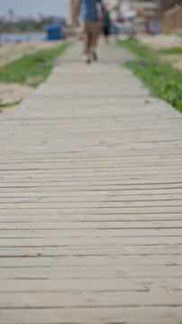 Man walking on beach boardwalk surrounded by greenery under sunny sky in summer coastal town setting with blurred background.