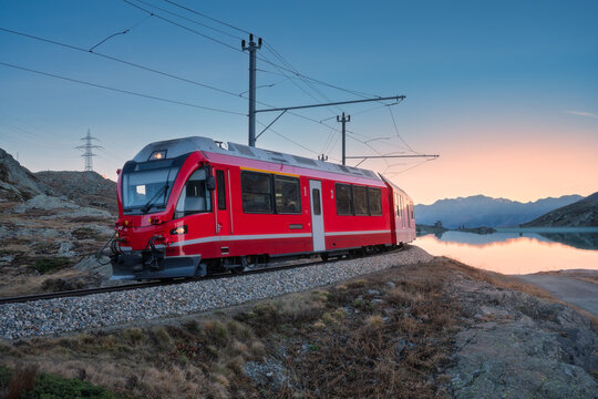 Fototapeta Bernina Express train near Lake Bianco in Switzerland at sunset, clear reflections, mountain silhouettes and alpine terrain in peaceful evening light in the Swiss Alps. Red train at dusk. Transport