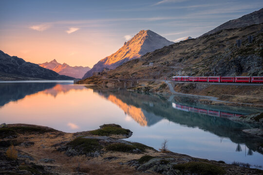Aerial view of Bernina Express train near Bianco lake in the Swiss Alps at sunset, with glowing mountain peaks, water with reflections, rocky shore. Peaceful autumn landscape. Mountain train in fall