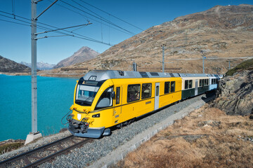 Obraz premium Yellow Bernina Express train traveling along lake Bianco in the Swiss Alps, Switzerland. Turquoise lake, rocky slopes, alpine mountains under bright blue sky on a clear autumn day. Glacier Express