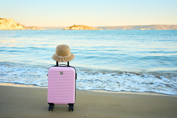 Pink suitcase with straw hat on sandy beach by the sea. Summer holiday, vacation and travel concept...