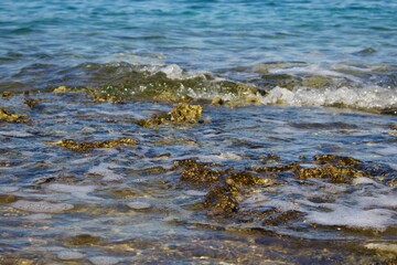  Clear sea water washes the rocky shore, creating small waves and bubbles of foam. Sunlight highlights the texture of the stones and the purity of the coastal waters.