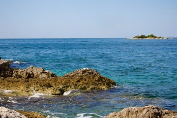 Landscape with sea and coastal rocks. The water shimmers with shades of blue and turquoise, and a small island with green vegetation is visible on the horizon.