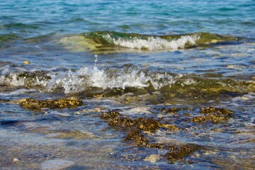 Transparent waves roll onto the rocky shore, forming white foam on the water surface. The clear sea shines in the sun, creating an atmosphere of freshness and tranquility.